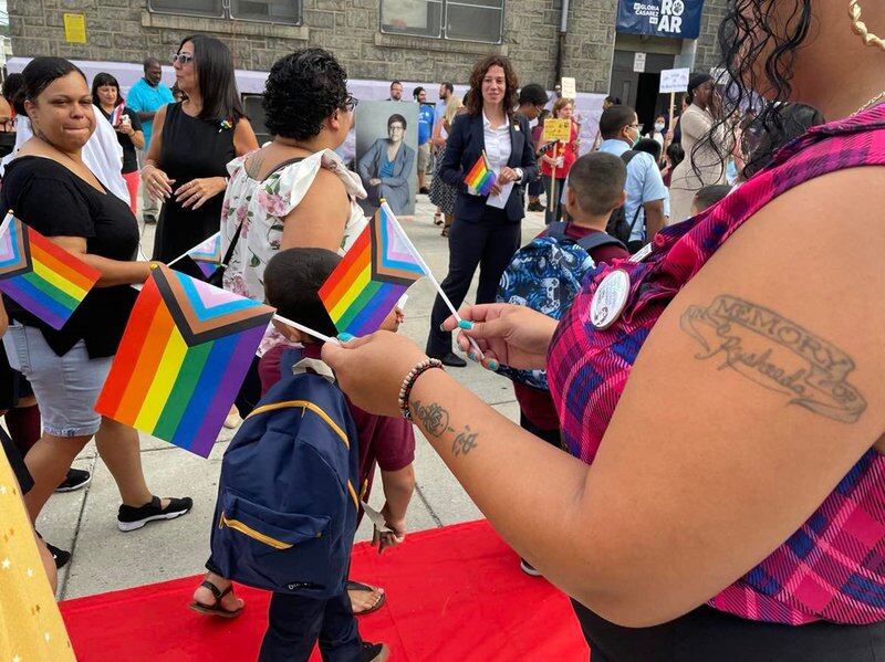 A small crowd of students and parents hold small pride flags inside an elementary school.