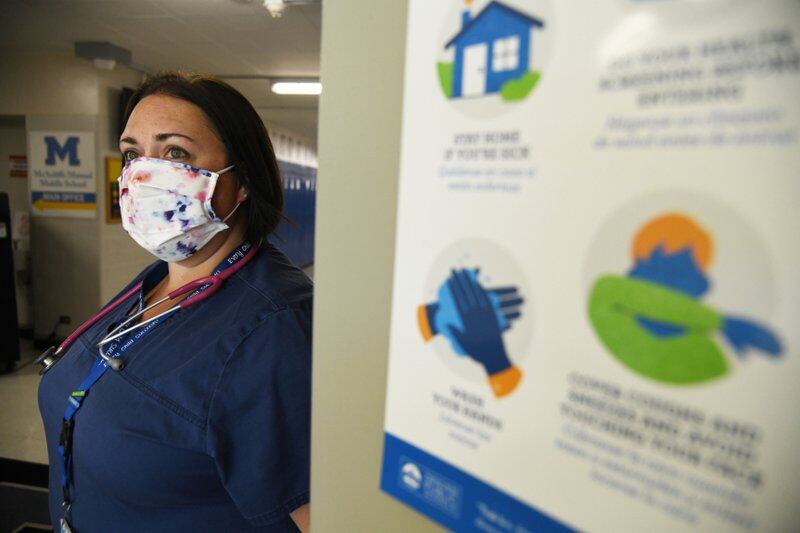 A school nurse leans agains a doorway. She is wearing blue scrubs and a face mask.