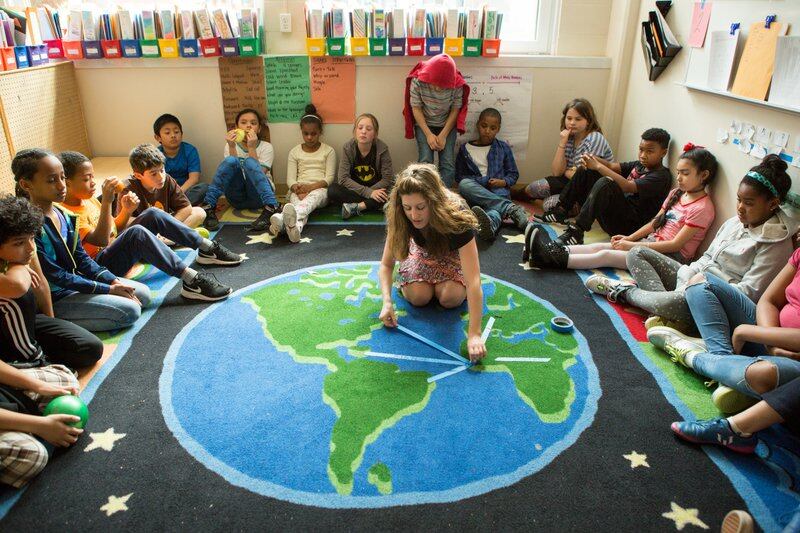 A teacher kneels on a rug shaped like the globe while students sit in a circle around her.