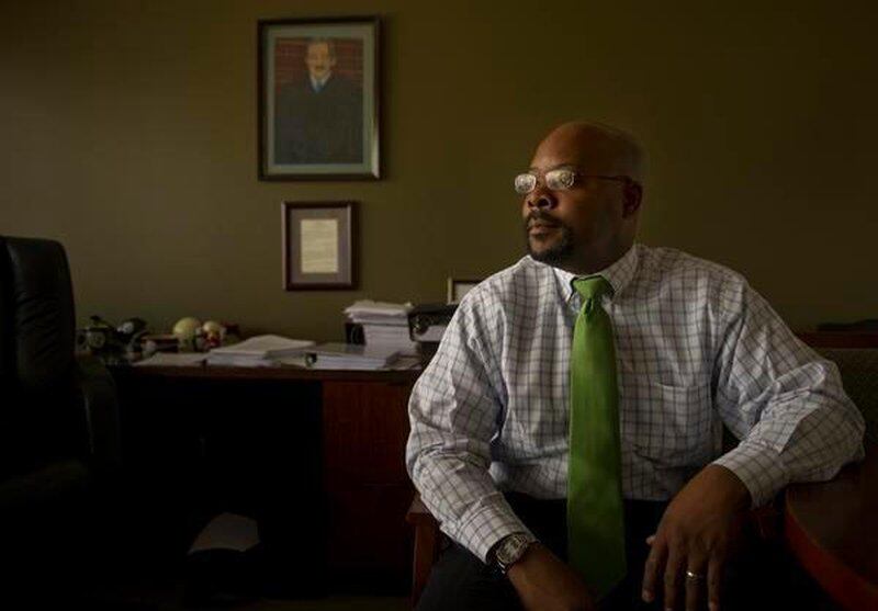 Aurora Superintendent Rico Munn, wearing a green tie and glasses, poses for a portrait while seated. A portrait of Supreme Court Justice Thurgood Marshall hangs on the wall.