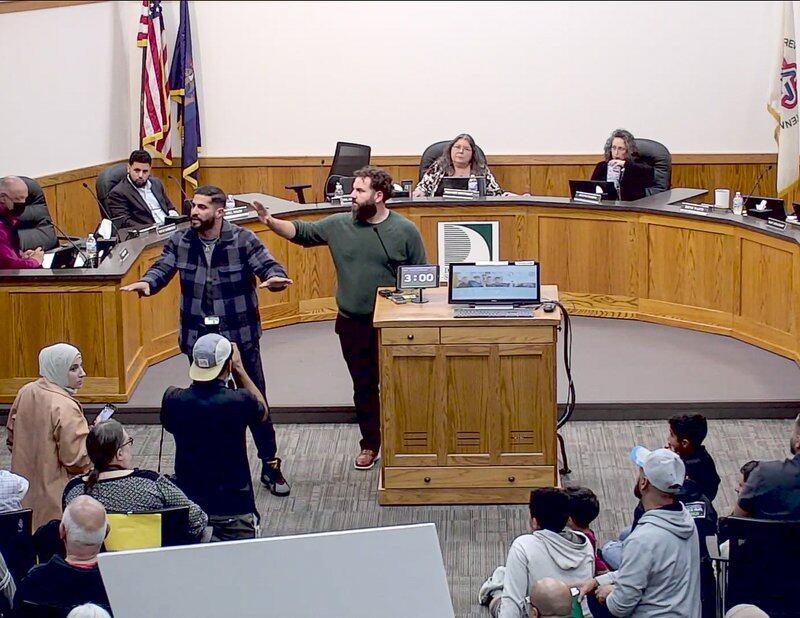 two men stand with arms up trying to calm a crowd in an administrative building as board members sit behind a semicircle desk in the background