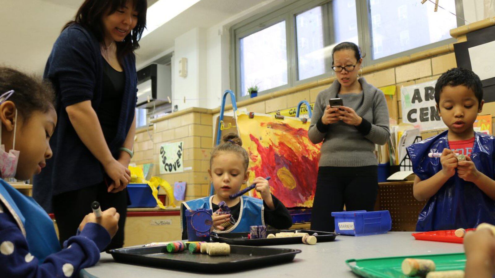 Yvette Ho, right, taps out a request to NYCHA to fix a leaky roof at CPC Jacob Riis Child Care Center. Meanwhile, a student shows her art project to Mary Cheng, who oversees early childhood programs for the Chinese-American Planning Council, a nonprofit that runs the daycare.