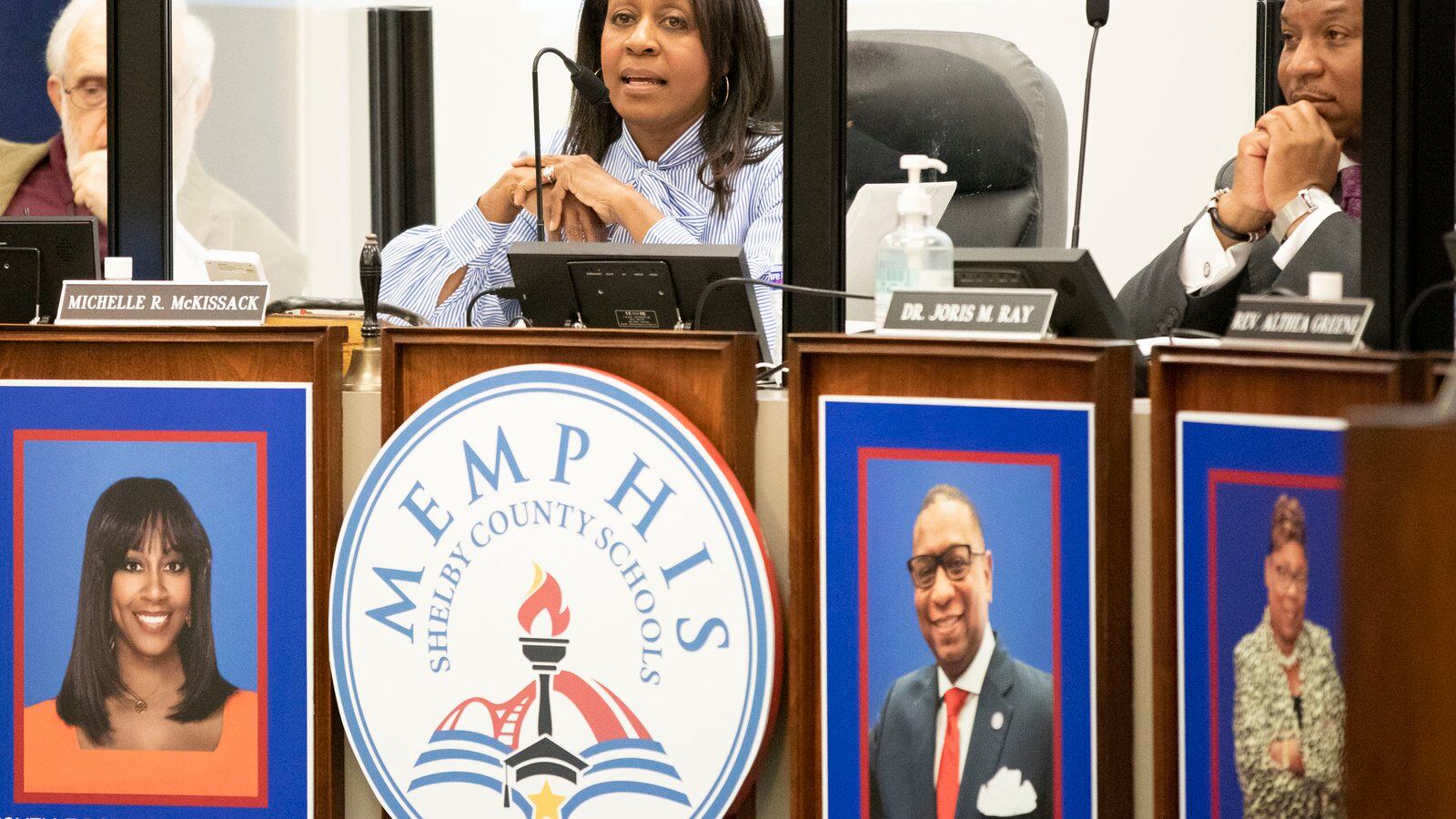 A woman sitting at a dais speaks into a microphone above a sign reading “Memphis Shelby County Schools”