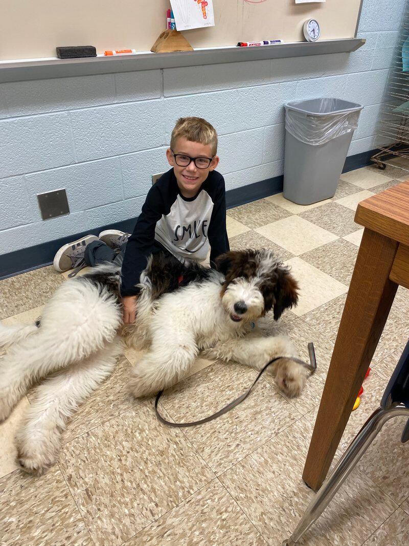 a student sits on the linoleum floor of a classroom with a hand on the dog sprawled in front of him