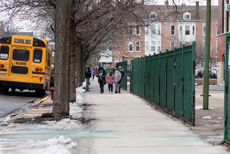 A man and two children walk down a sidewalk by a yellow school bus.