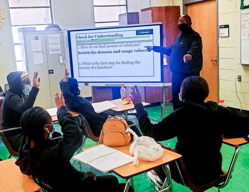 A male teacher stands in a classroom in front of a large monitor as engaged students watch from their desks and raise their hands to contribute to the discussion. 