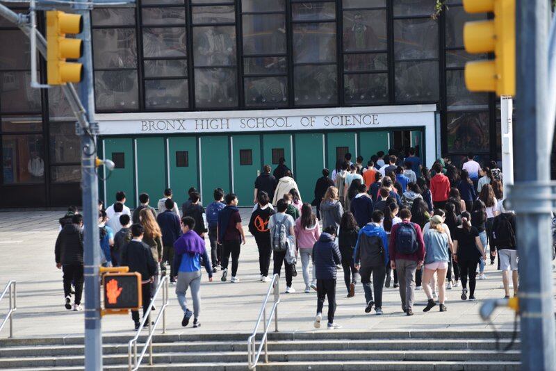 Several students walk toward the entrance of Bronx High School of Science in New York City. They are framed by two yellow traffic lights.