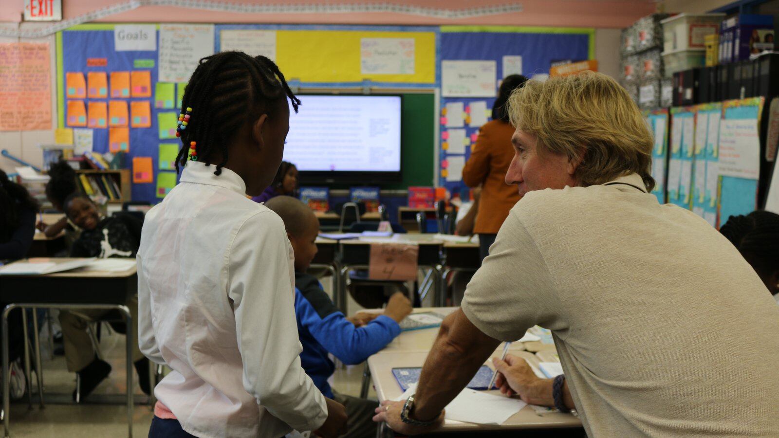 A teacher works at a desk with a young student with braided hair wearing a white dress shirt in the classroom.
