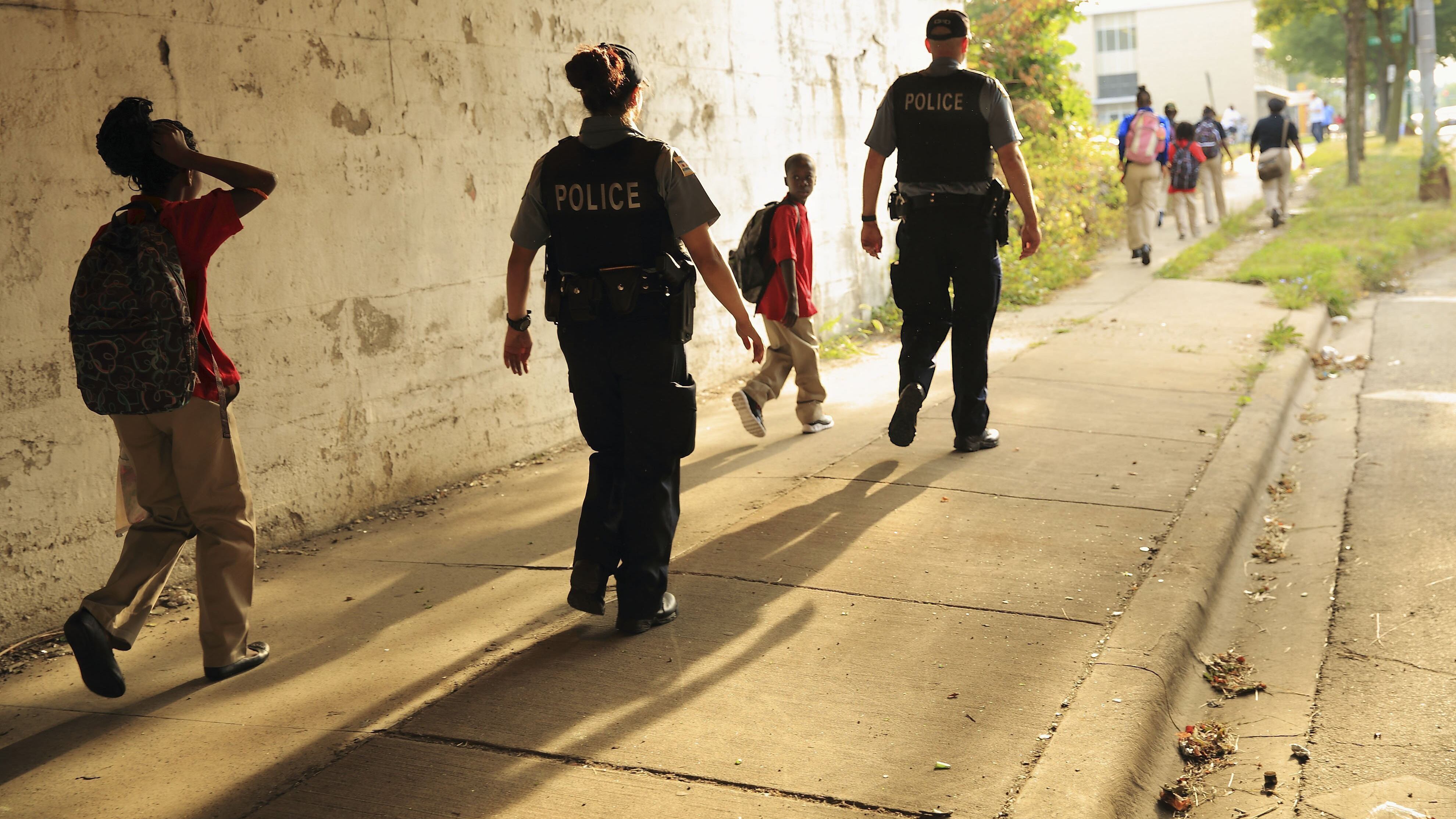 Two Chicago Police Department officers, wearing black vests with “POLICE” in white lettering on the back, escort children to school. The sun casts long shadows behind those walking down the sidewalk.