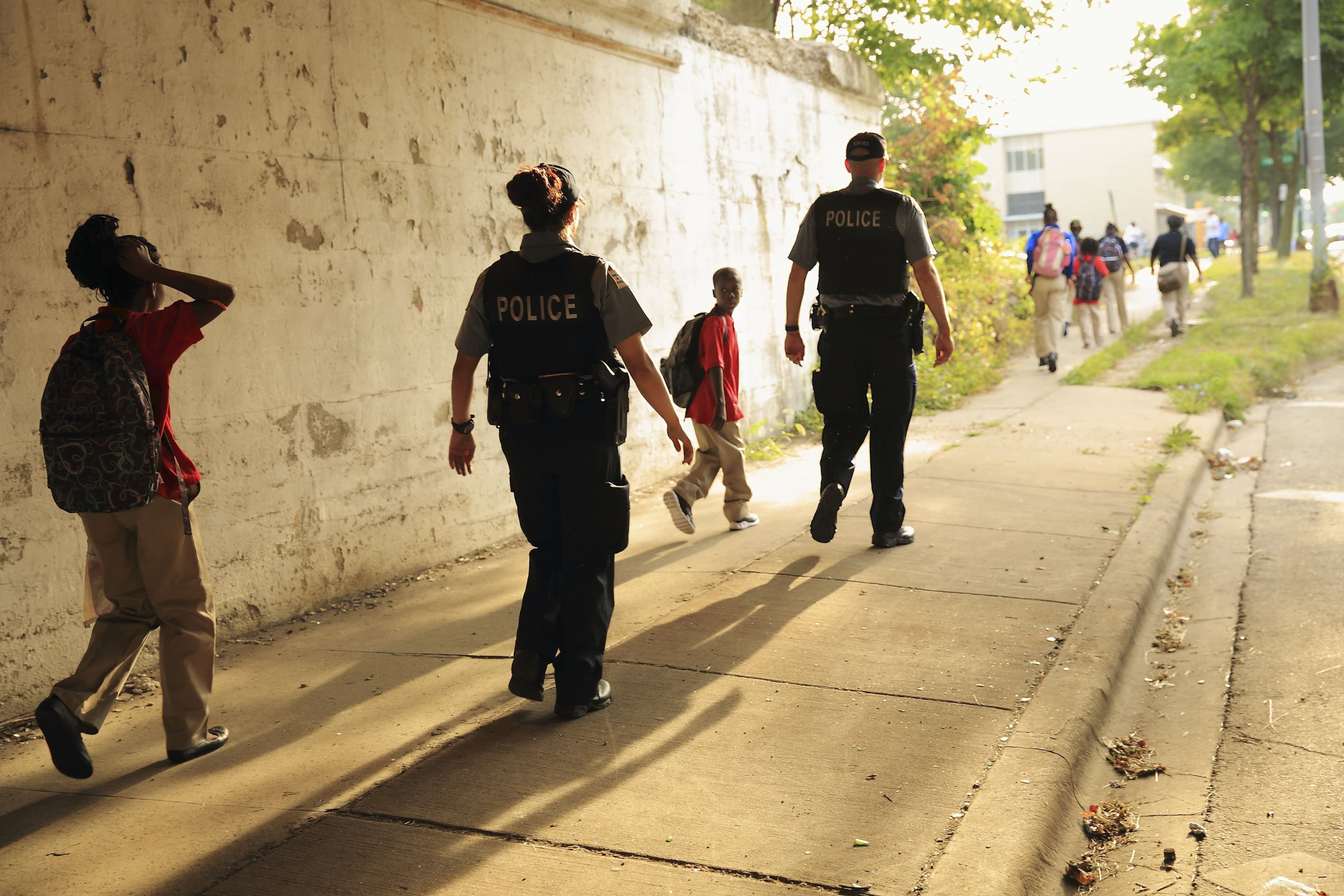 Two Chicago Police Department officers, wearing black vests with “POLICE” in white lettering on the back, escort children to school. The sun casts long shadows behind those walking down the sidewalk.