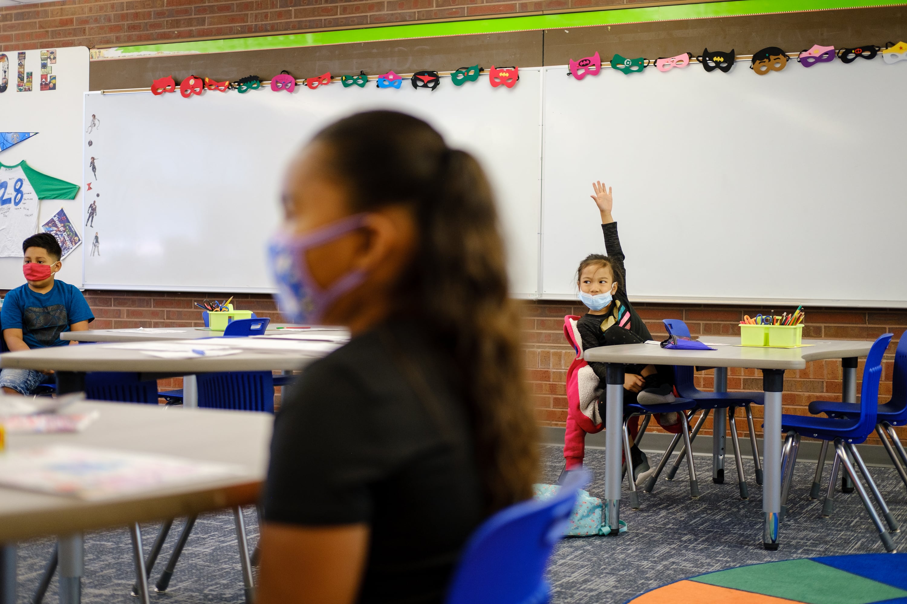 A young student at a table in a classroom raises his hand to speak. Another student in the foreground is slightly blurred. Both students are wearing masks.
