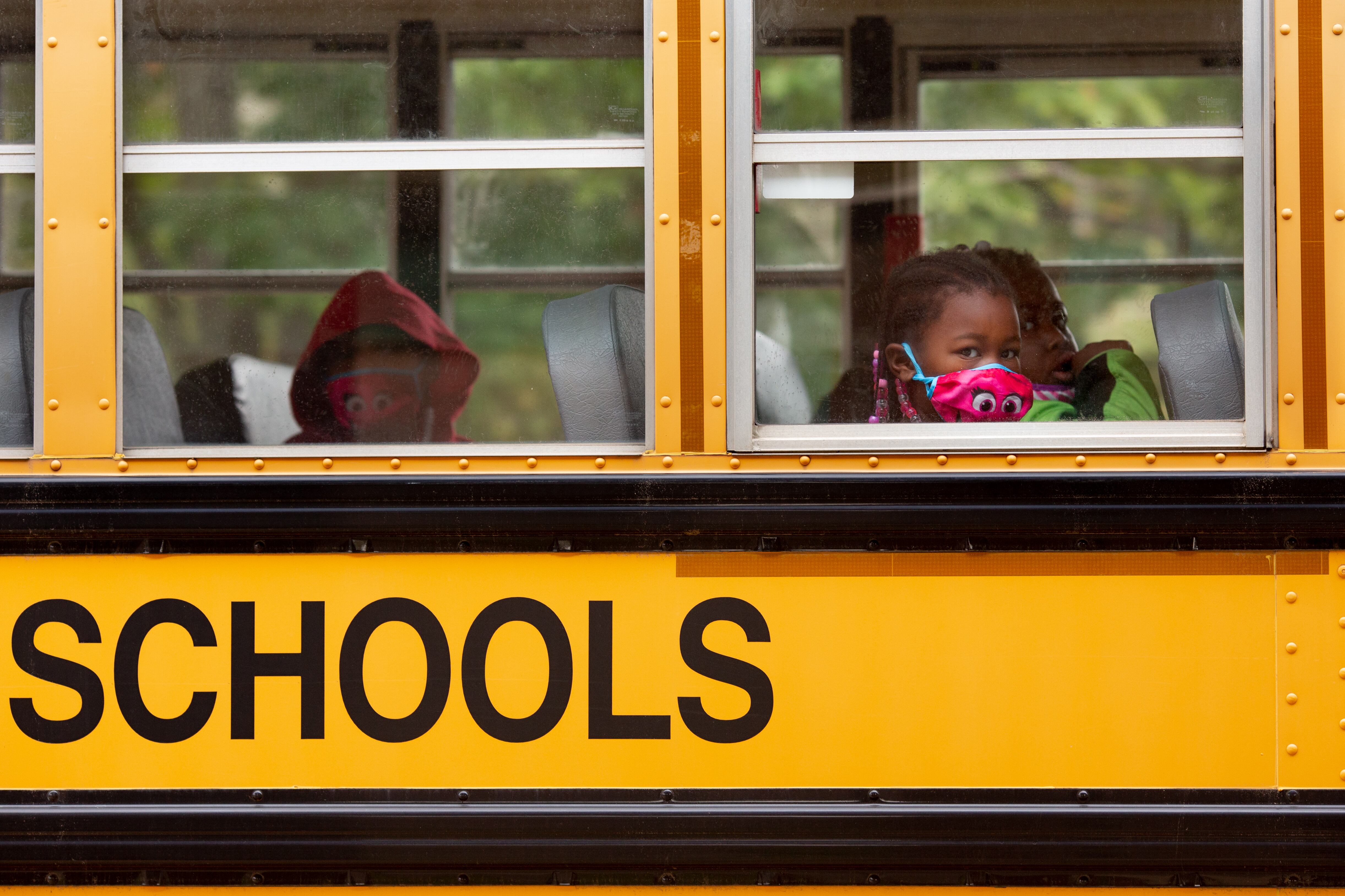 Close-up from outside a yellow school bus shows three young students wearing colorful face masks sitting in two rows.