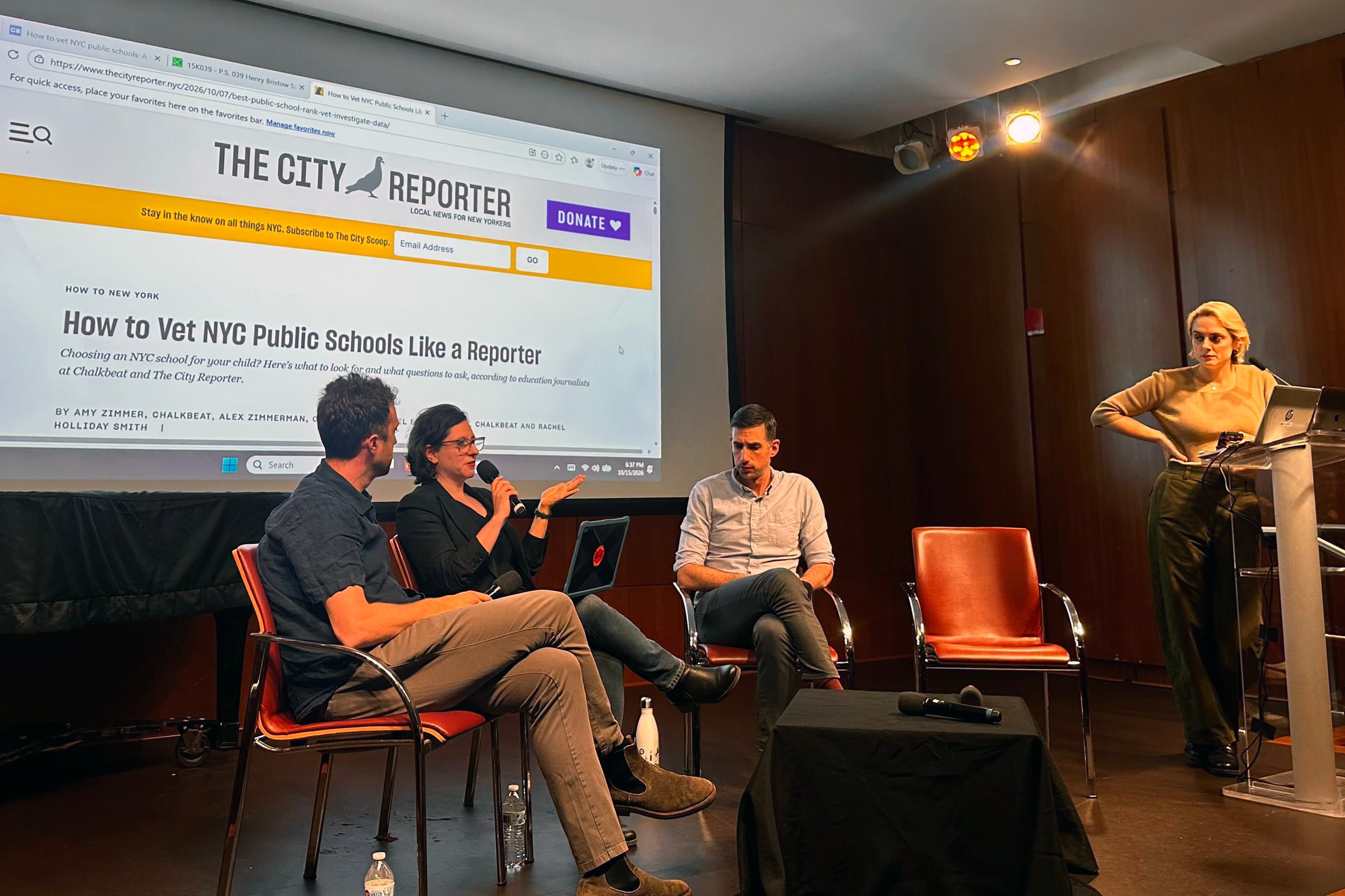 Four adults on stage with a large screen in the background showing a journalism story.
