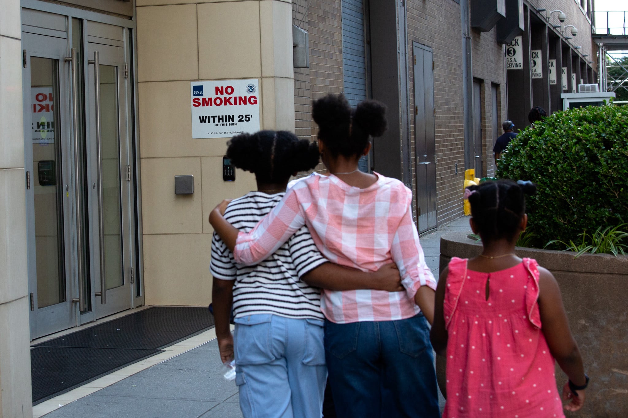 A view from behind three young Black children embrace each other while they walk past a federal building.
