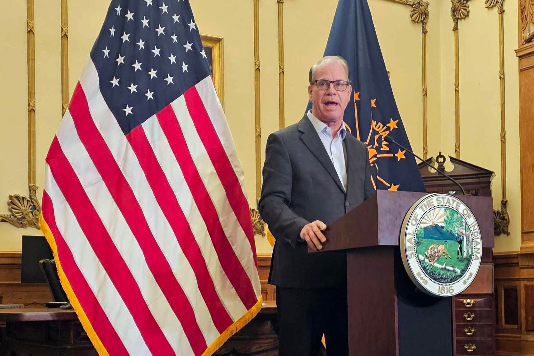 A man stands behind a podium and in front of an American and an Indiana state flag.