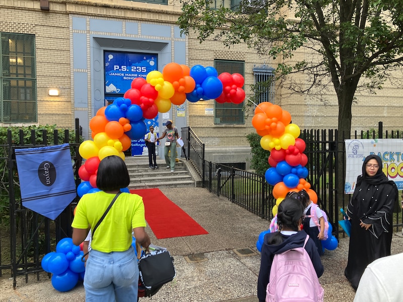 Adults and students stand outside the front entrance to a school building with a colorful balloon arch.