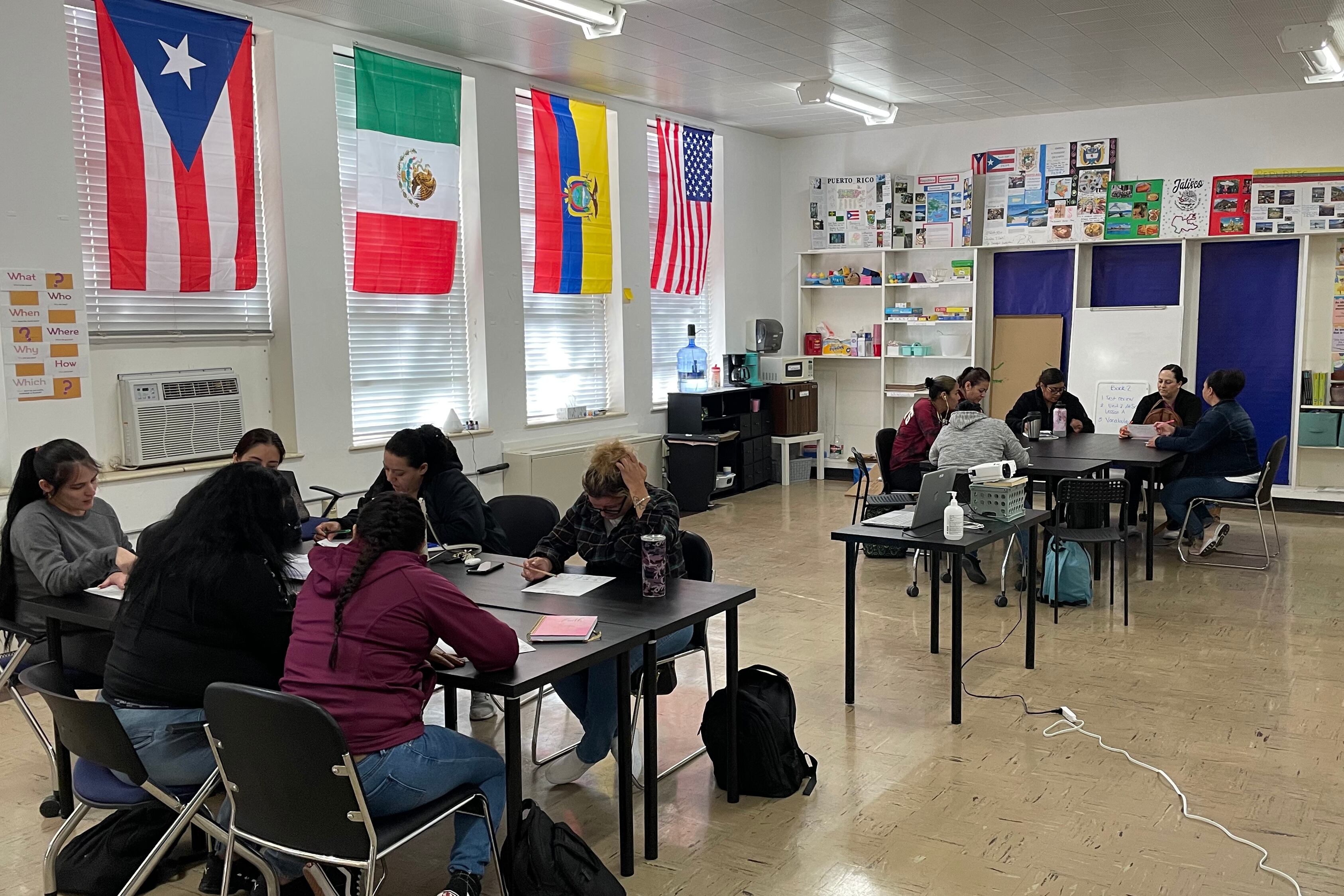 Two large tables full of adult students in a classroom with flags from different places along the wall.