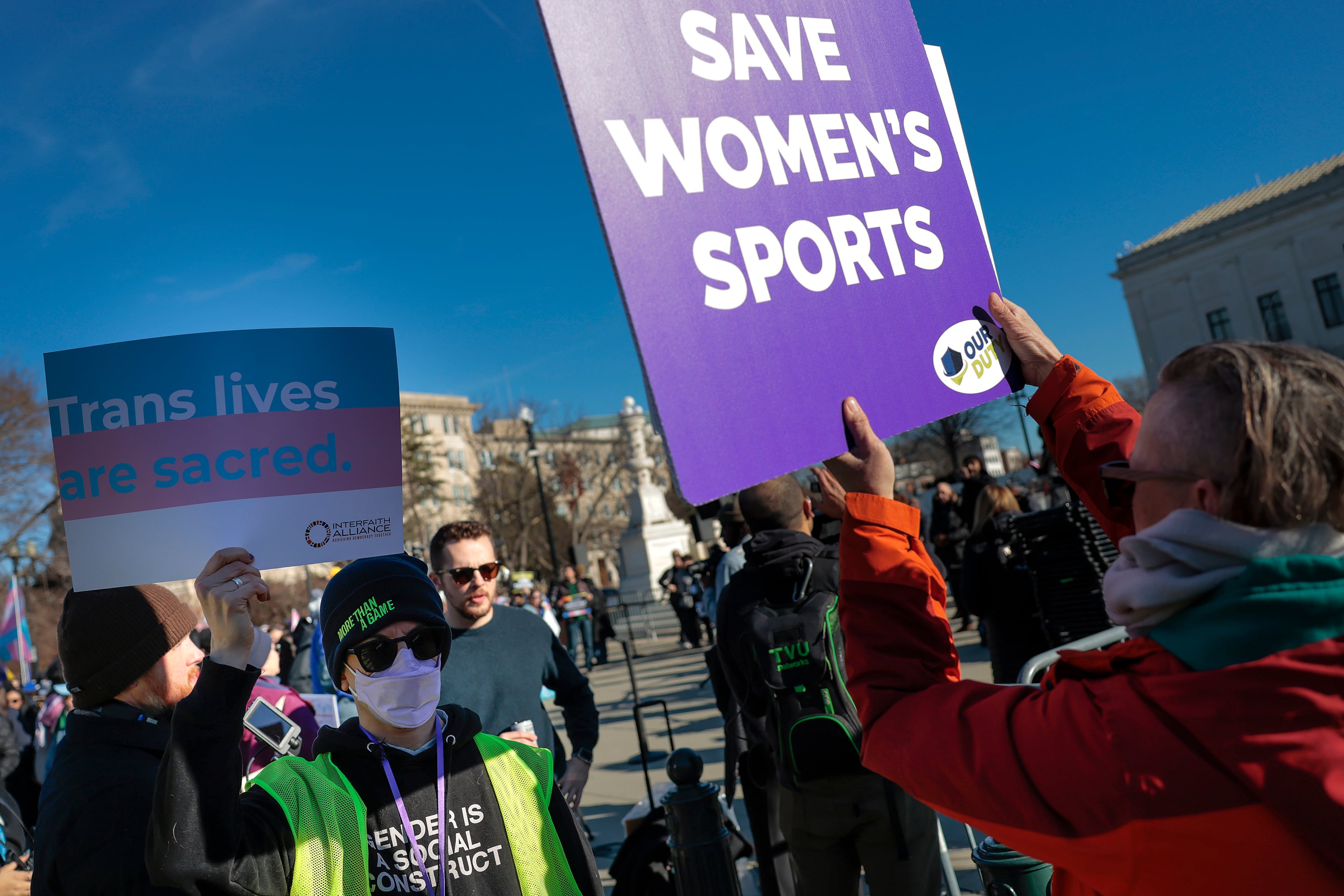 A photograph of protesters wearing sunglasses and masks holding signs that read"Save Women's Sports" and "Trans lives are sacred."