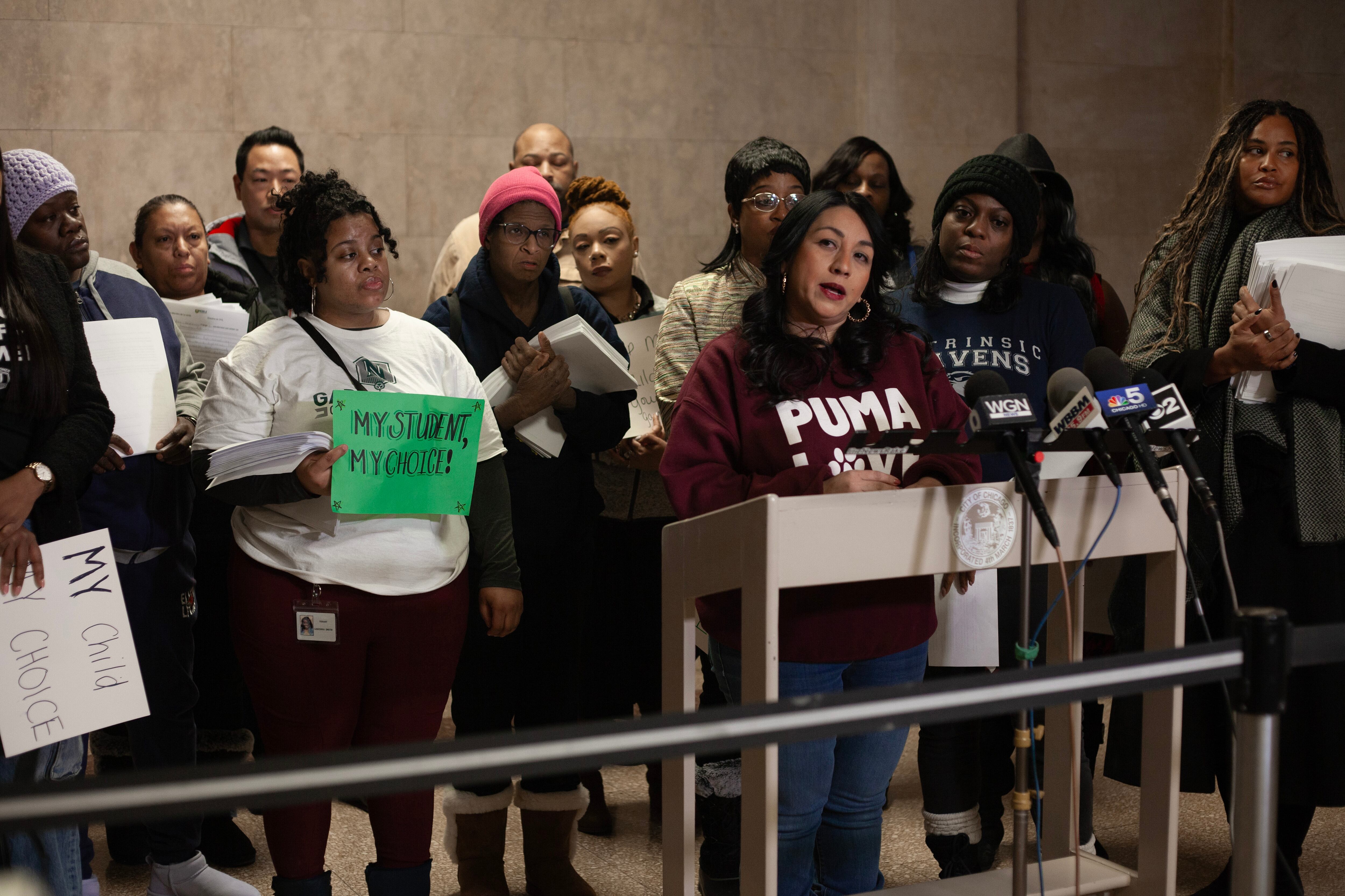 An adult with long dark hair and wearing a red sweater stands at a podium with microphones while a group of people holding signs stand in the background.