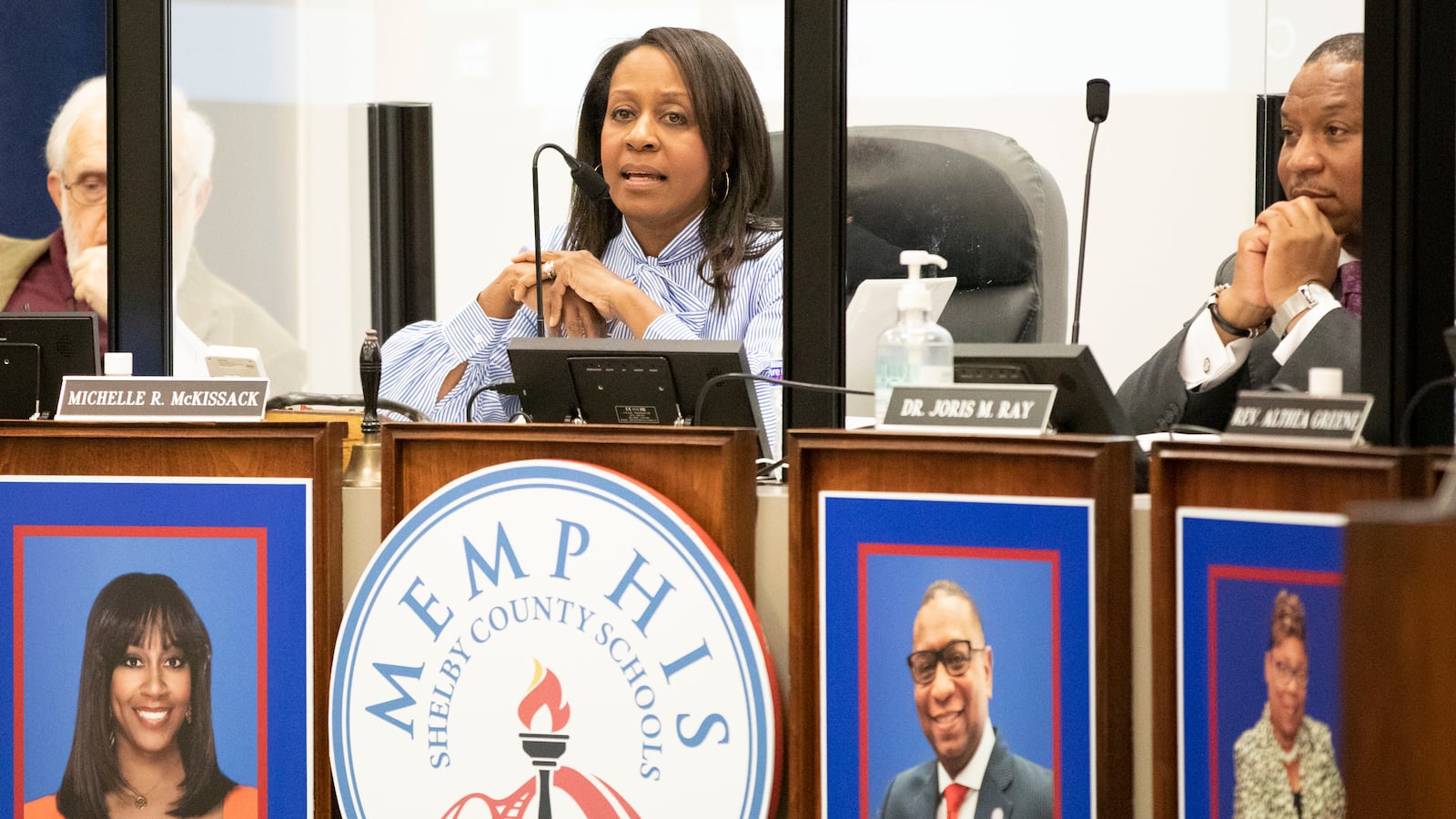 A woman sitting at a dais speaks into a microphone above a sign reading “Memphis Shelby County Schools”