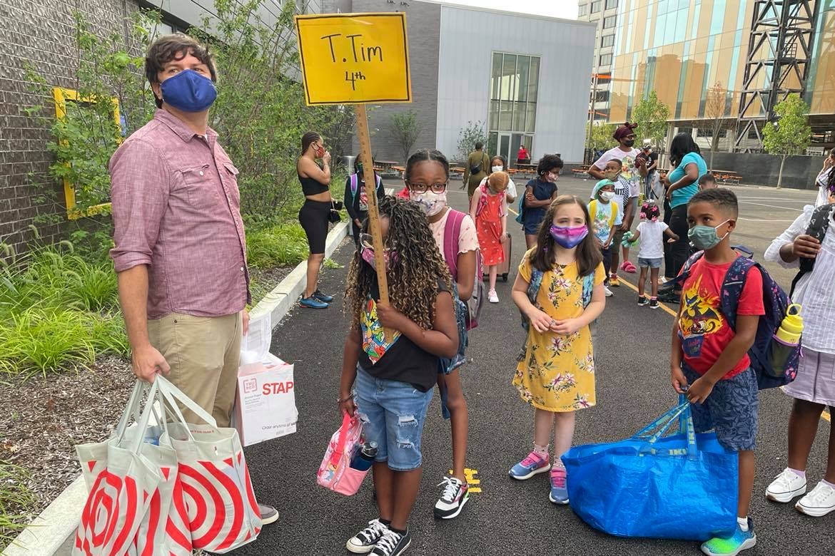 A man in a light red shirt holds Target bags and stands next to his students outside a school.