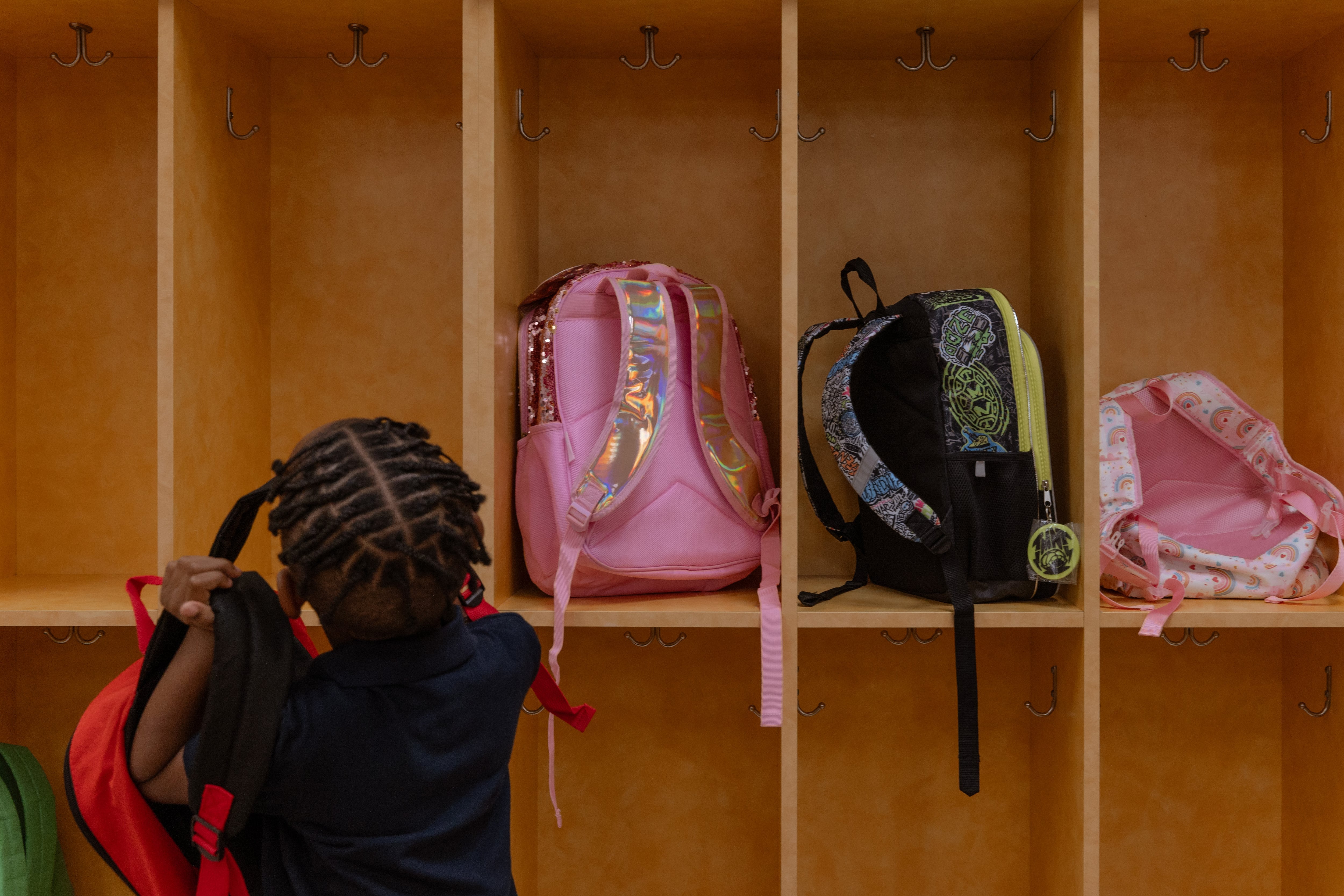 A view of the back of a student putting their backpack away next to other backpacks on a wooden cubby.
