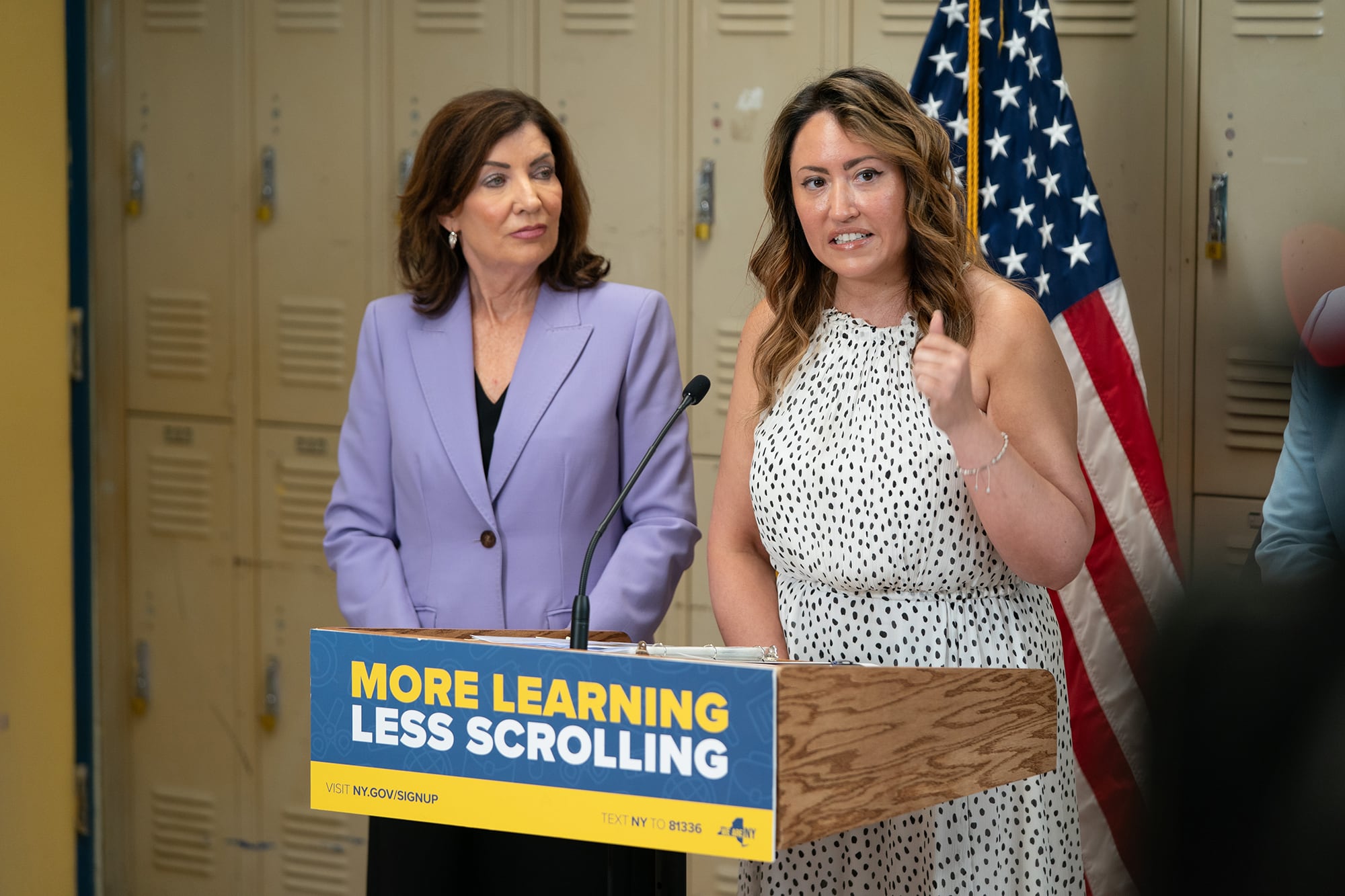 Two women in business clothes stand behind a wooden podium in a school hallway with lockers and an American flag in the background.