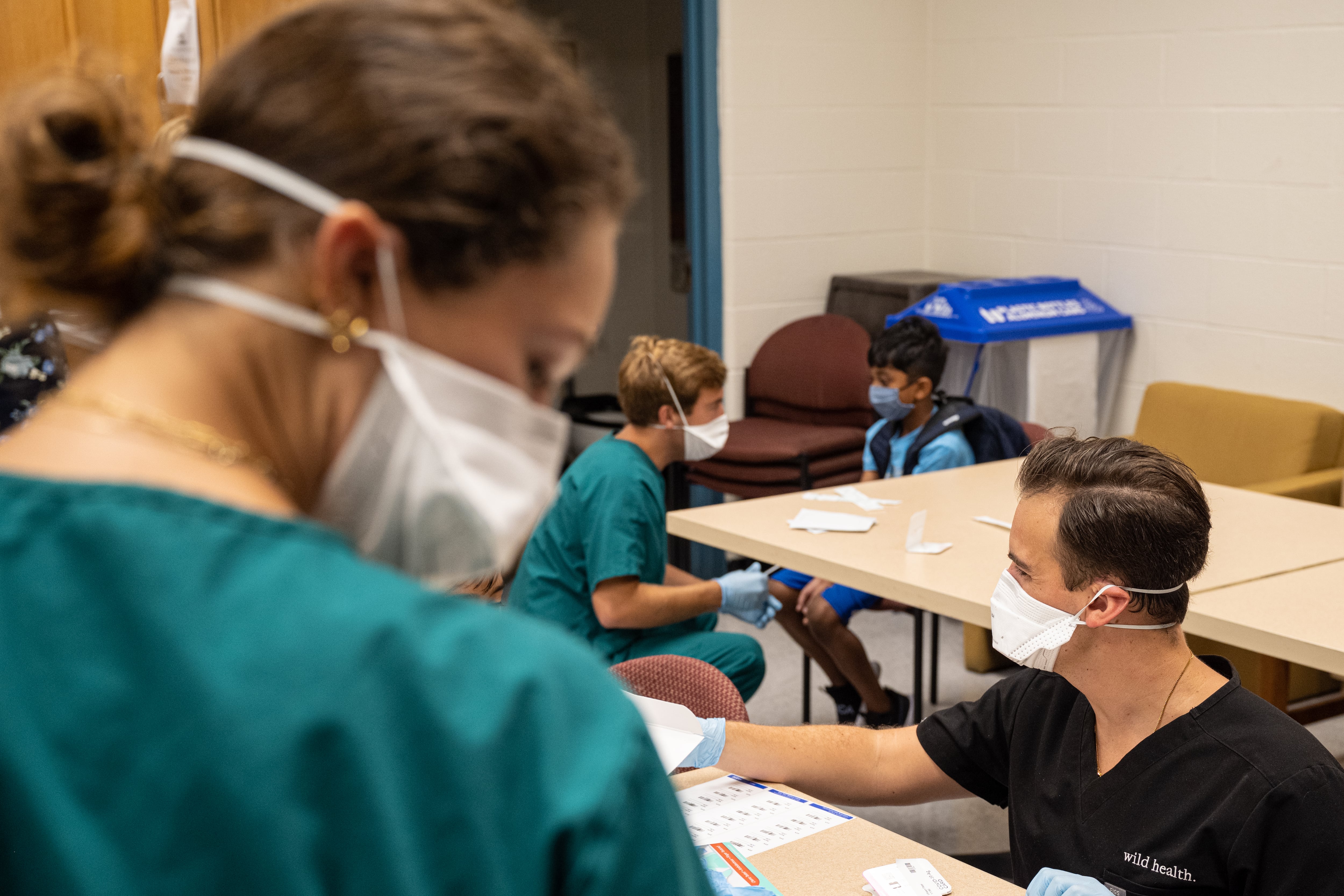 A medical professional administers a COVID test to a young boy. In the foreground of the photo is a close up of a medical professional with a white mask and a green scrub top.