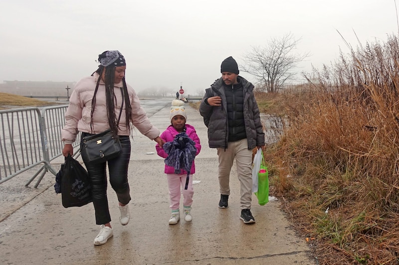 Two adults and a young child wearing cold weather clothing walk toward the camera on a sidewalk outside. A grey sky in the background.
