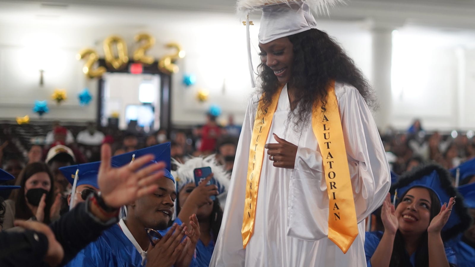 A young woman, wearing a white cap and gown with a yellow salutatorian sash, stands to give her address to classmates and family members.
