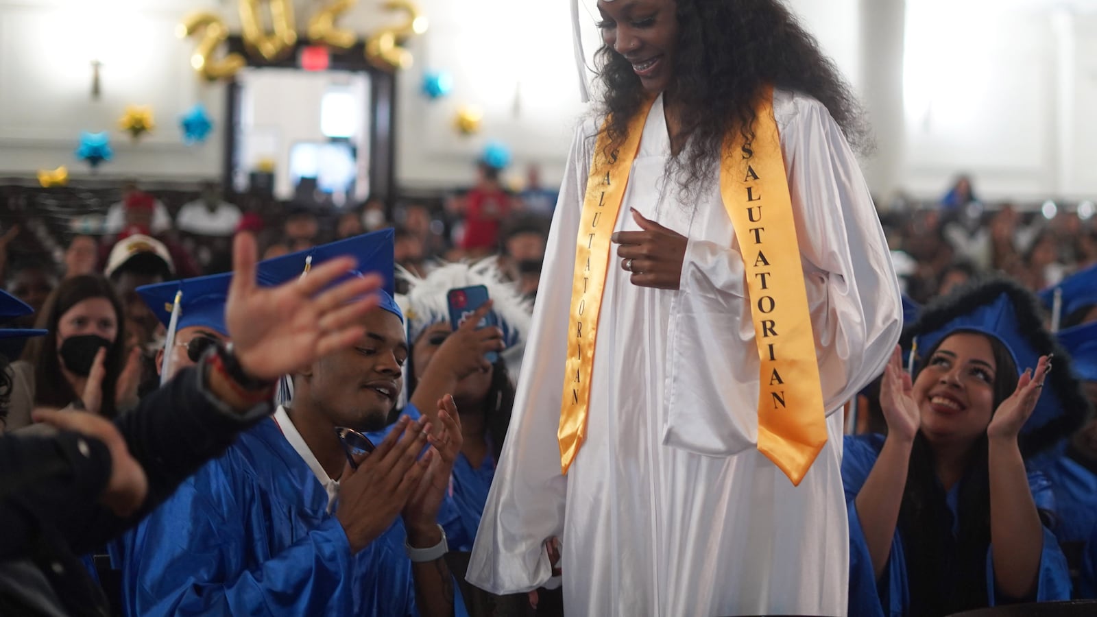 A young woman, wearing a white cap and gown with a yellow salutatorian sash, stands to give her address to classmates and family members.