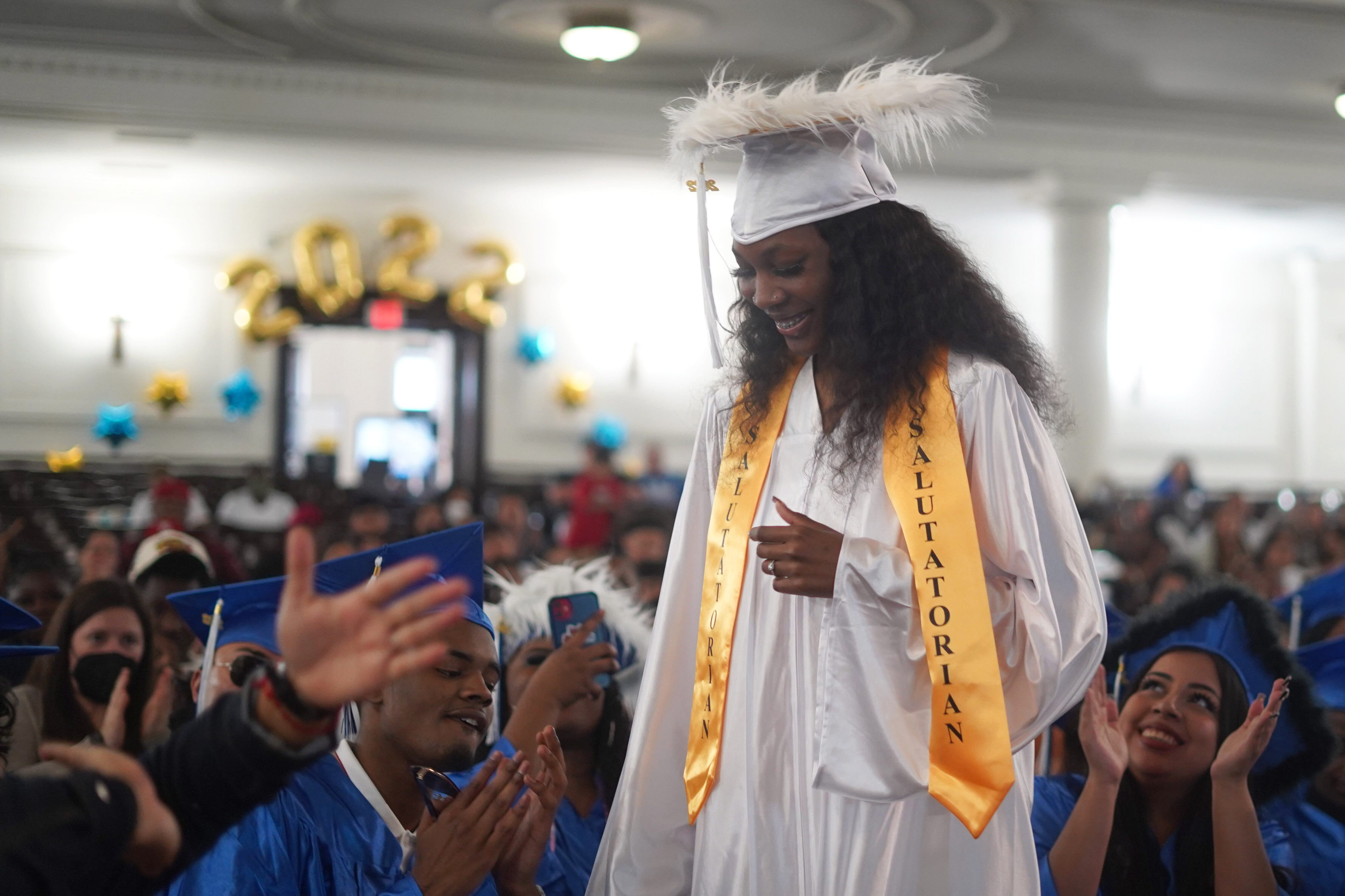 A young woman, wearing a white cap and gown with a yellow salutatorian sash, stands to give her address to classmates and family members.