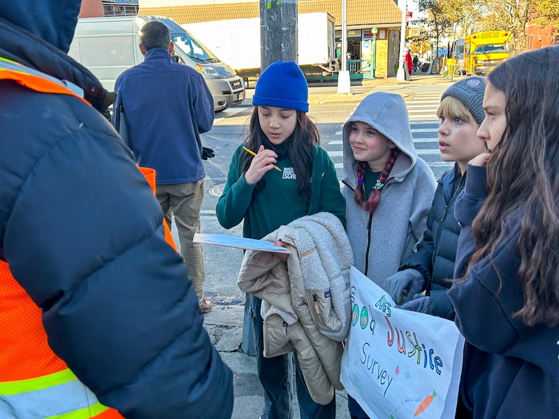 Four young students stand with coats and hats on outside while they talk to an adult.