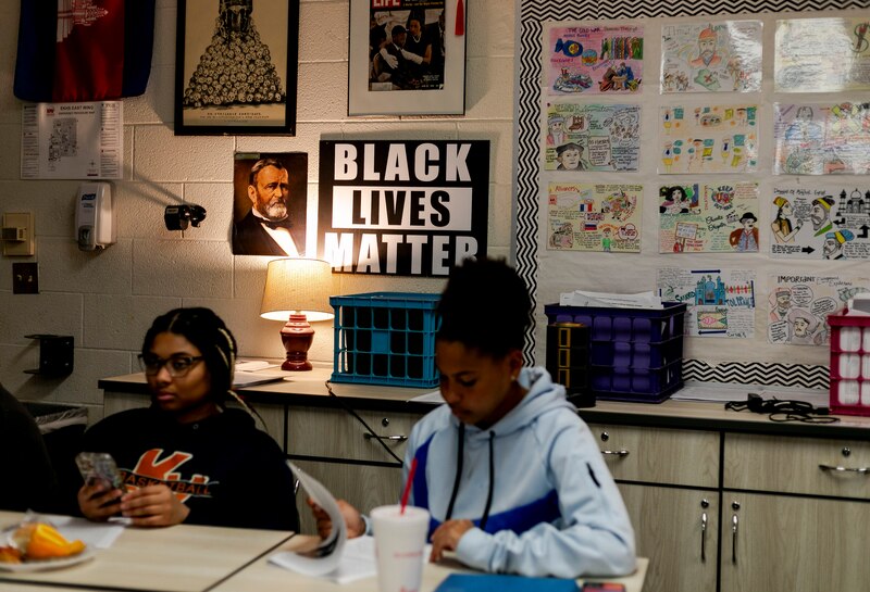 Two high school students work from a desk in a classroom with a wall full of posters in the background.