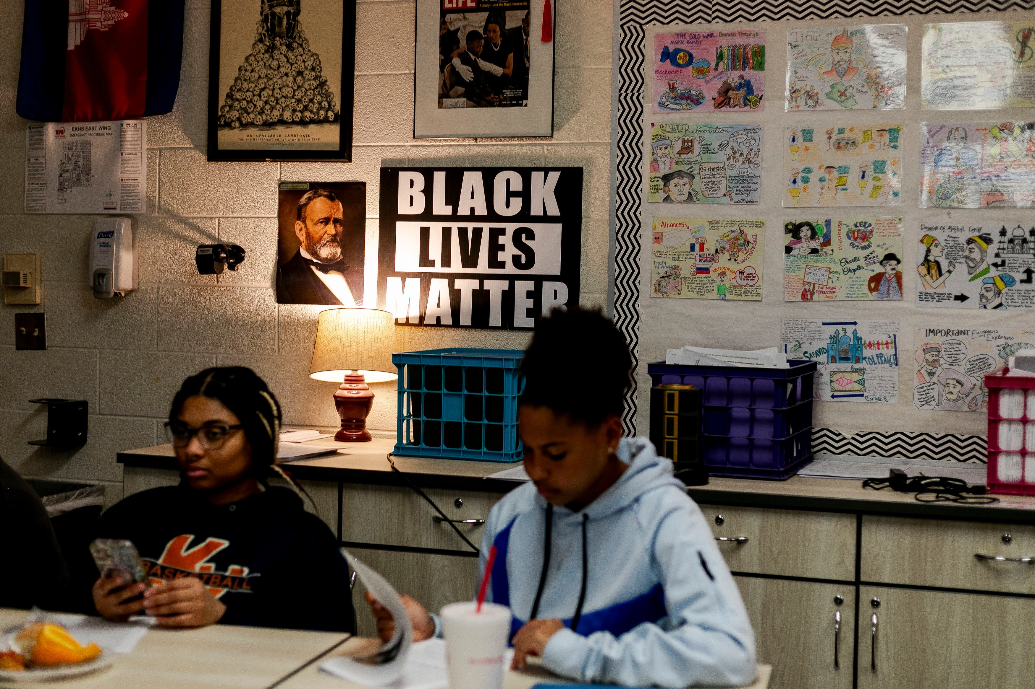 Two high school students work from a desk in a classroom with a wall full of posters in the background.