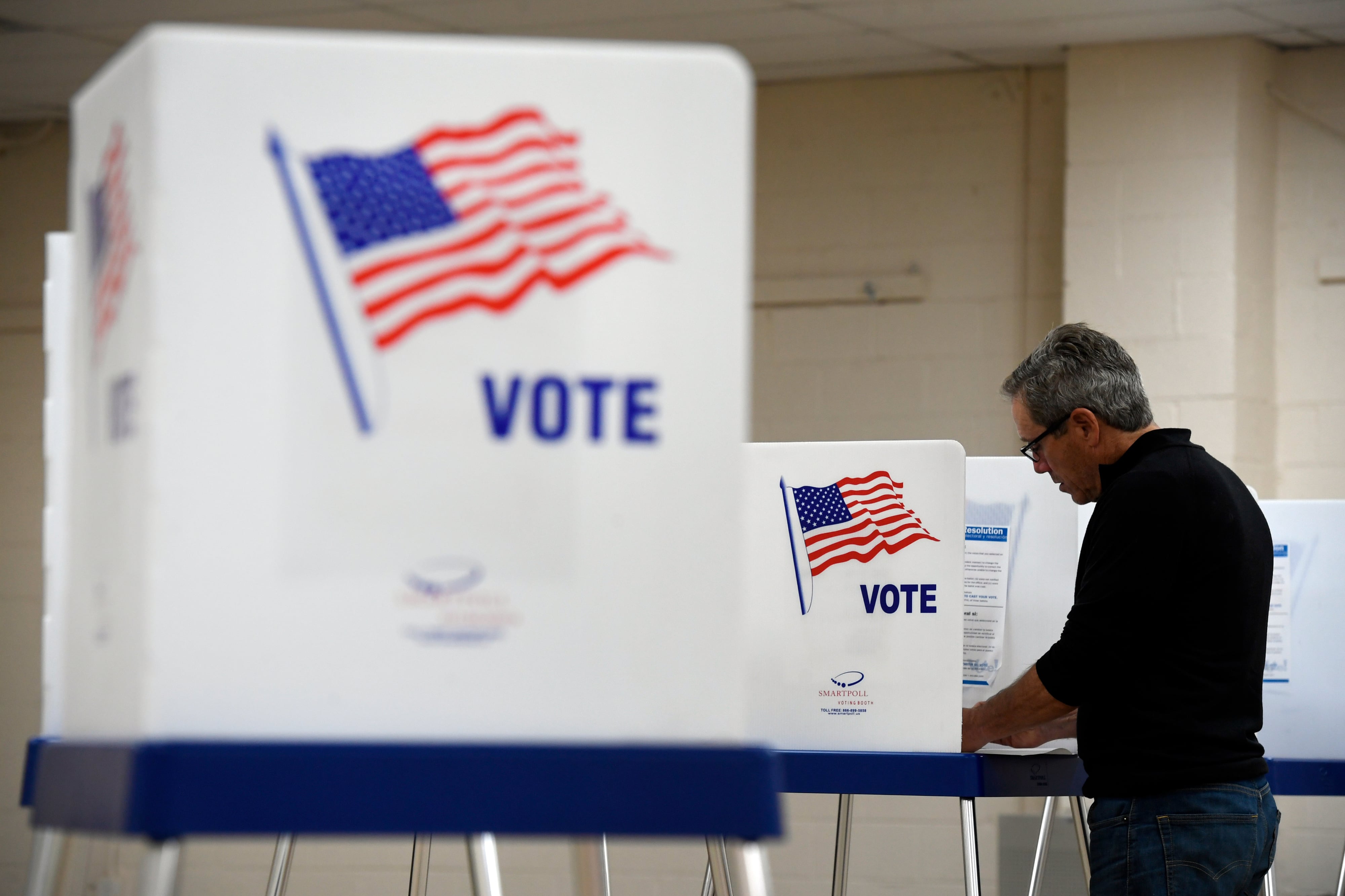 A man goes over his ballot in a voting booth, with a few other white booths sit in the foreground. Each voting partition is decorated with an American flag and the word “VOTE”.