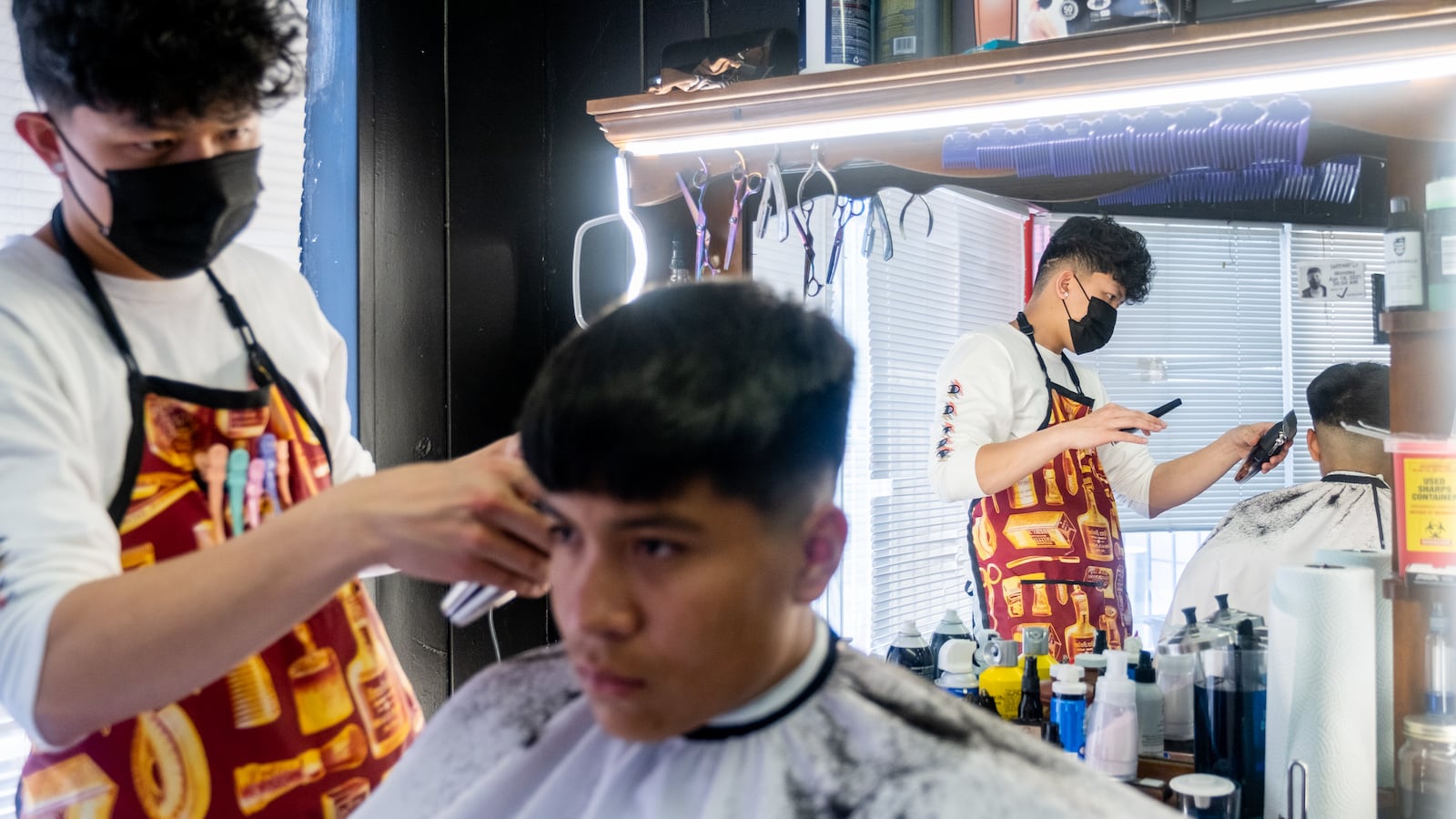 A young man wearing a black mask and barber’s apron cuts the hair of another young man in a barber shop. The barber is reflected in his mirror, adorned with bright lights at the top.