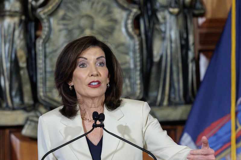 New York Gov. Kathy Hochul speaks at a press conference. She has dark hair and is wearing a white suit. She stands behind a podium and gestures with one hand.