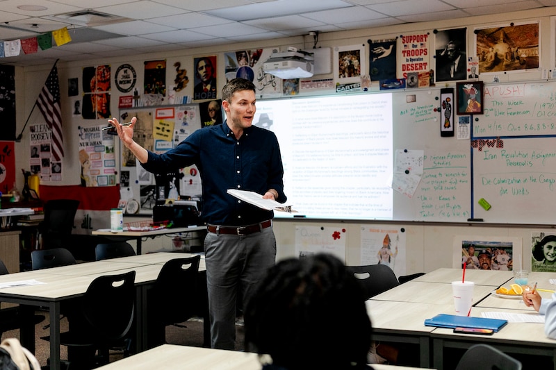 A high school teacher wearing a dark button up shirt speaks to a class of high school students with a large projector and a decorated wall in the background.
