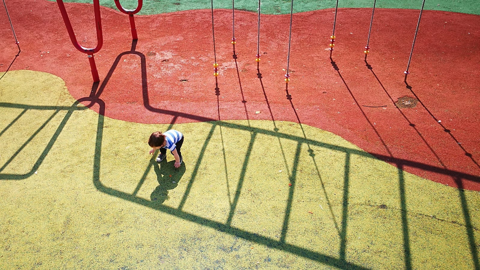 A small boy leans down to touch his shadow on a red and yellow playground surface.