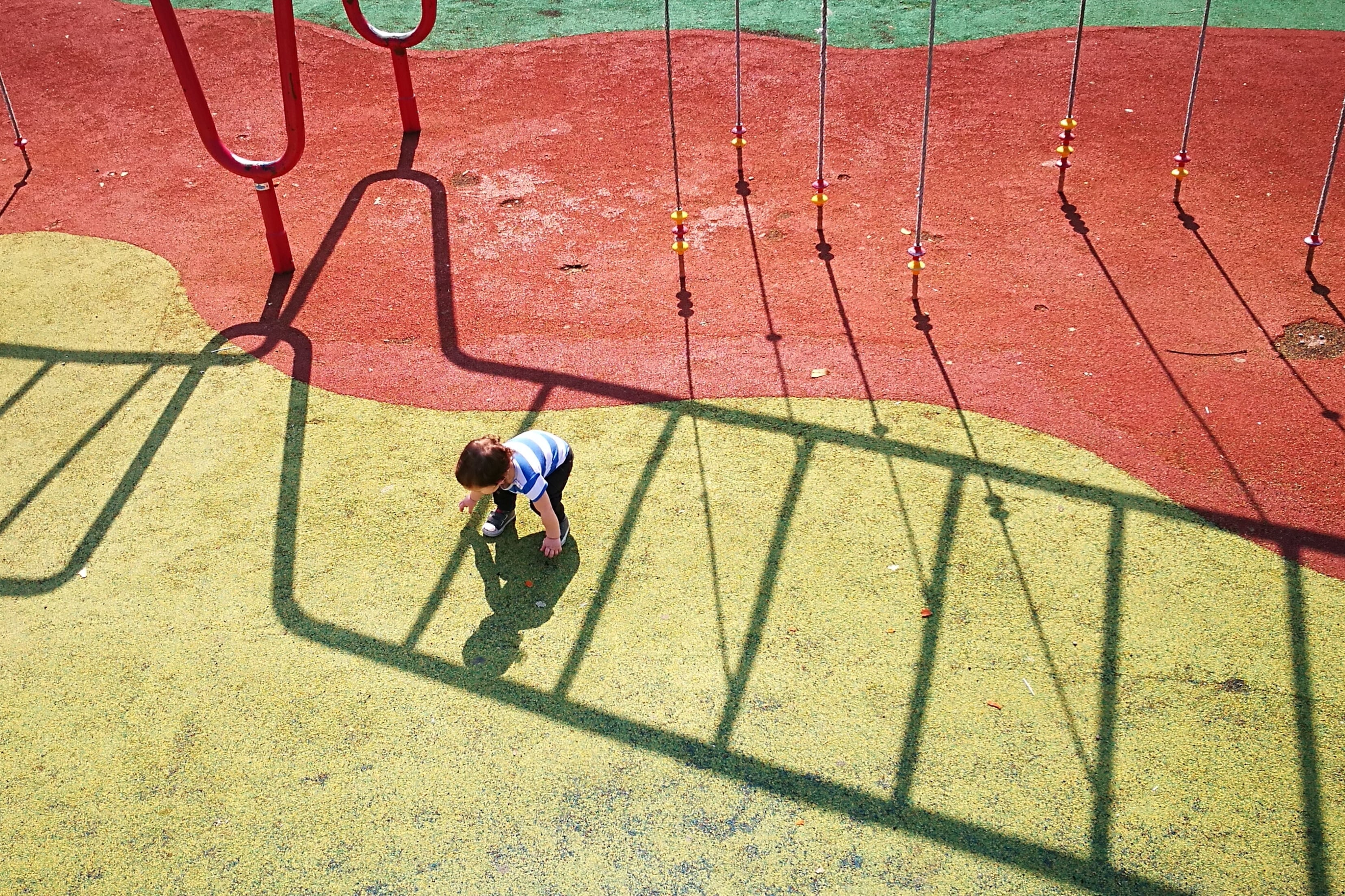 A small boy leans down to touch his shadow on a red and yellow playground surface.