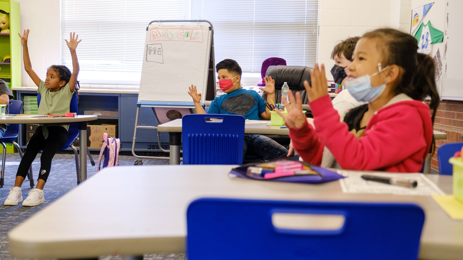 Elementary students excitedly raise their hands together at their desks. Some students are wearing protective masks, while others are not.