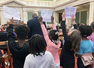 Adults and students gather together in a corner of a large meeting room, some are holding signs.