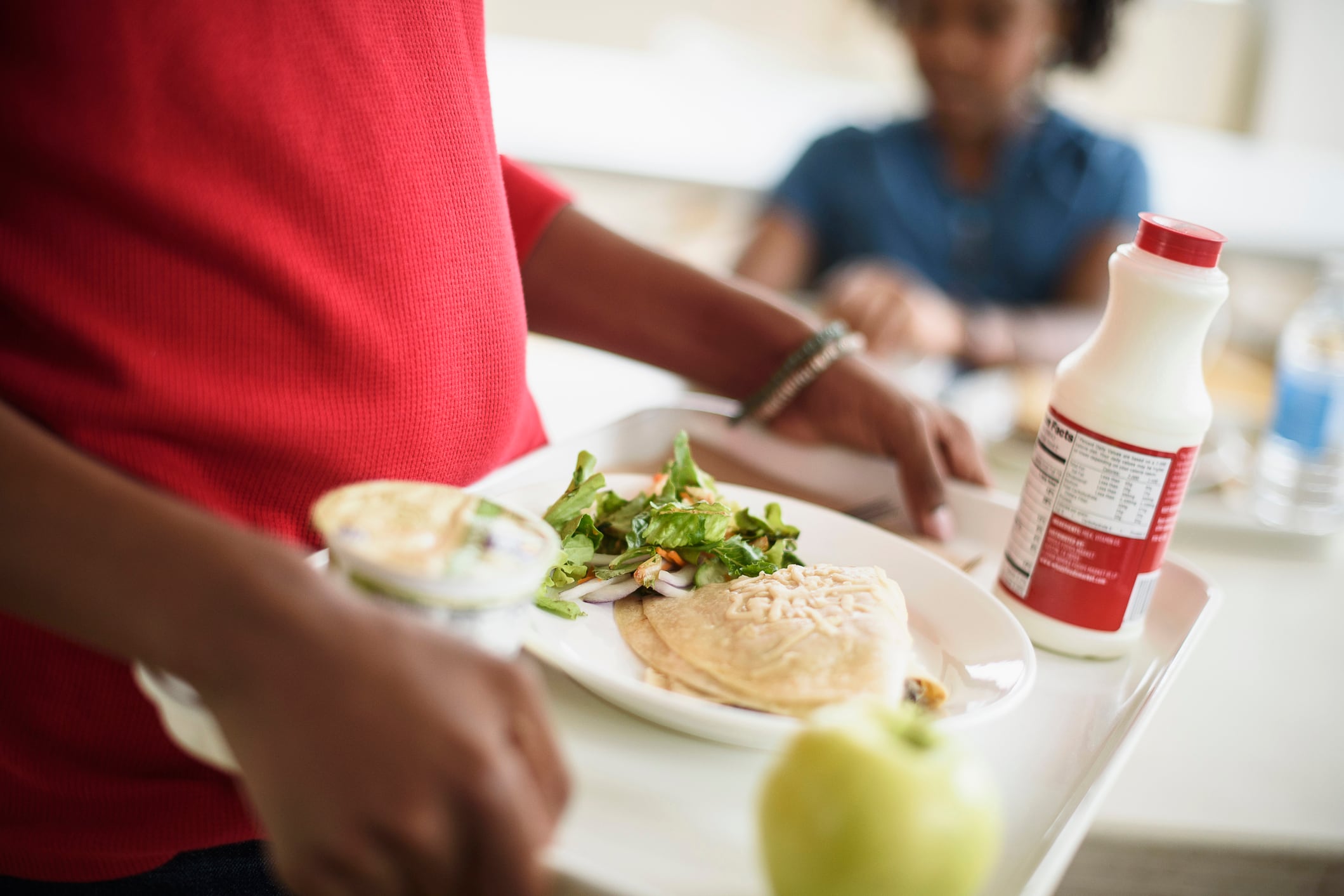A close up of a young student carrying a tray full of school food including milk, an apple, a salad and other things. A student sitting at a table is in the background.