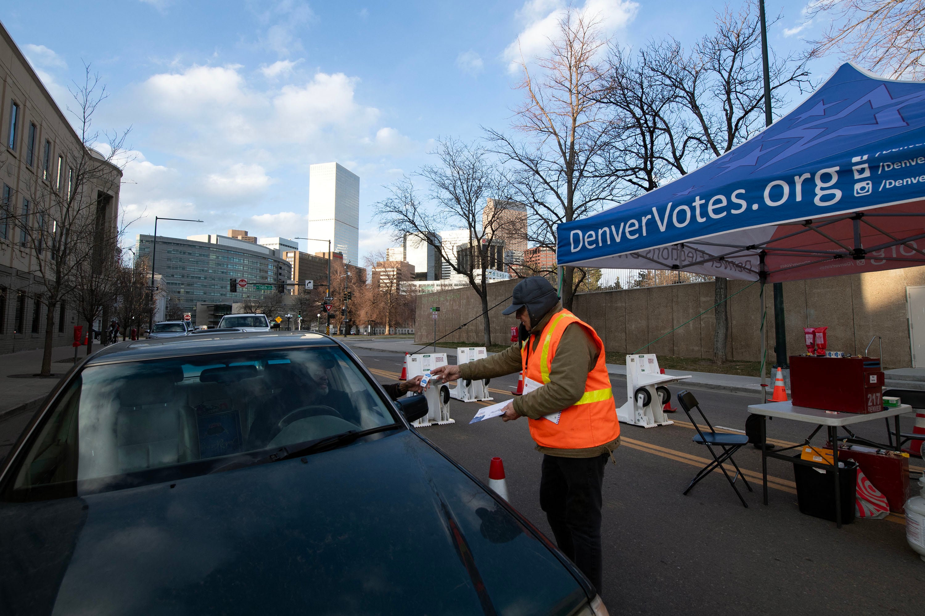 A person wearing a safety vest grabs a mail-in ballot from a person sitting in a car on a street with buildings and a blue and cloudy sky in the background.