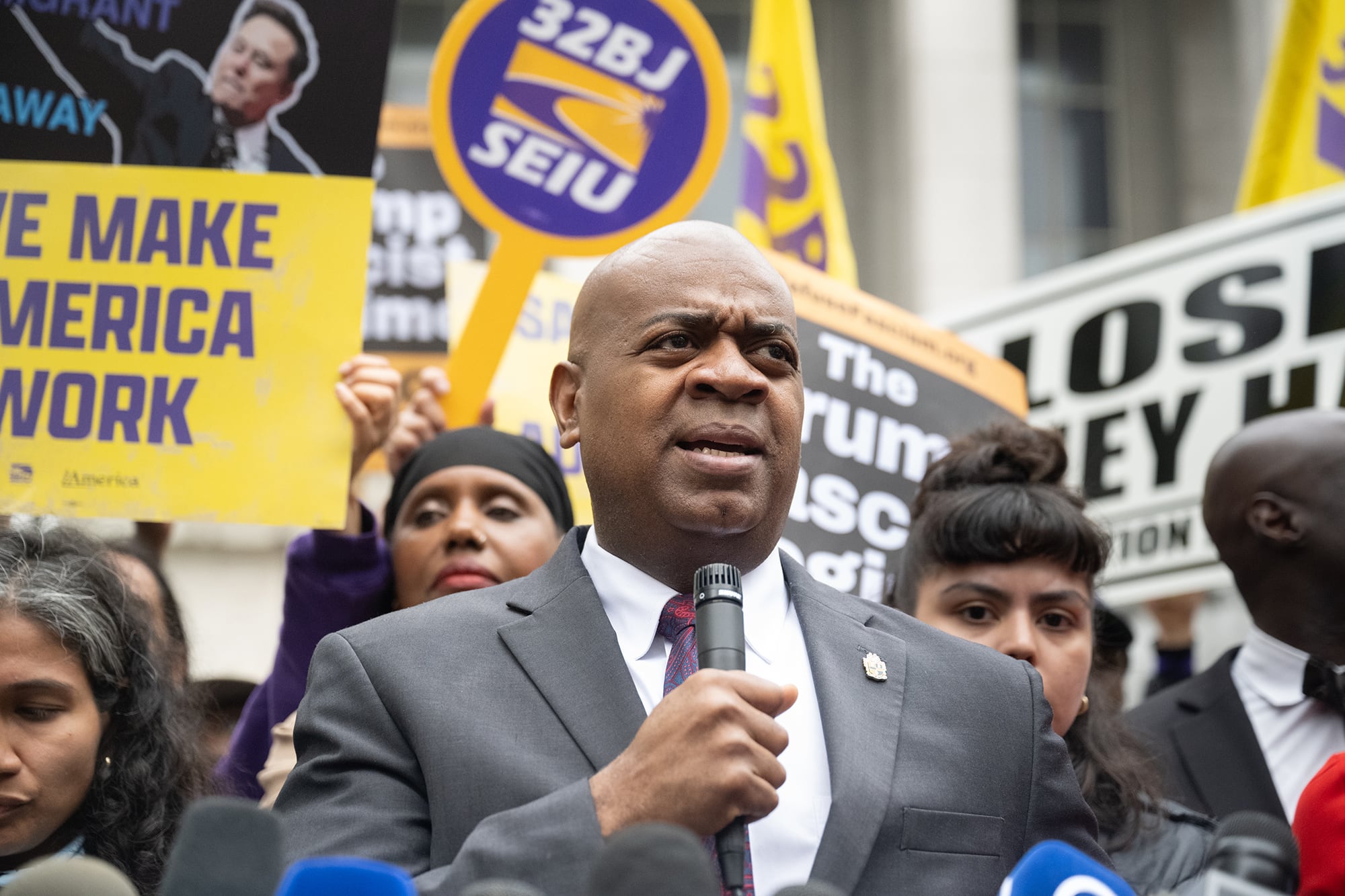 A Black man in a grey suit and holding a microphone speaks in front of a large crowd outside. Some people are holding large signs.