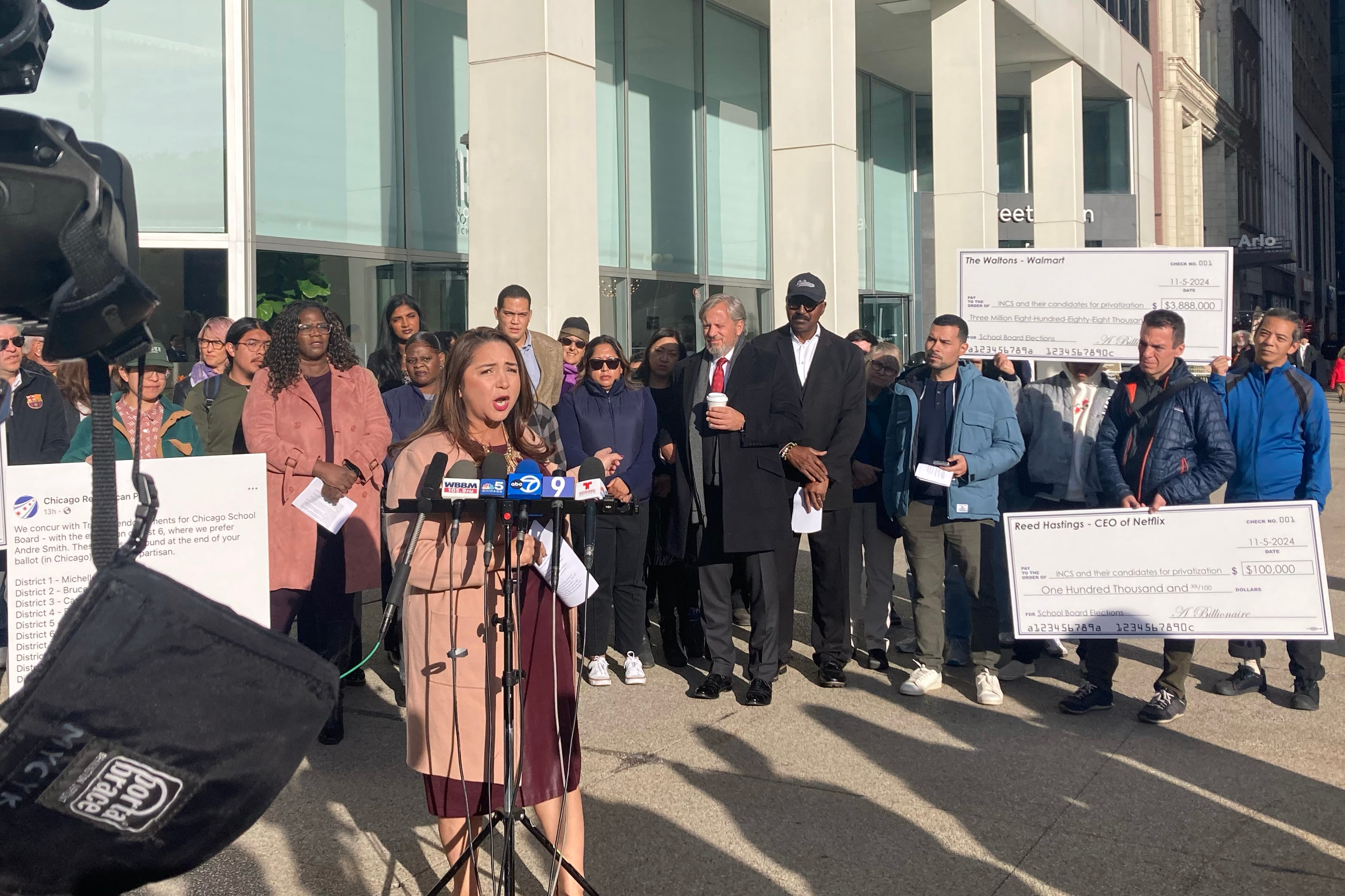 A group of people stand outside, some are holding giant checks and posters while one woman in a pink coat speaks from behind a podium with a cluster of microphones.