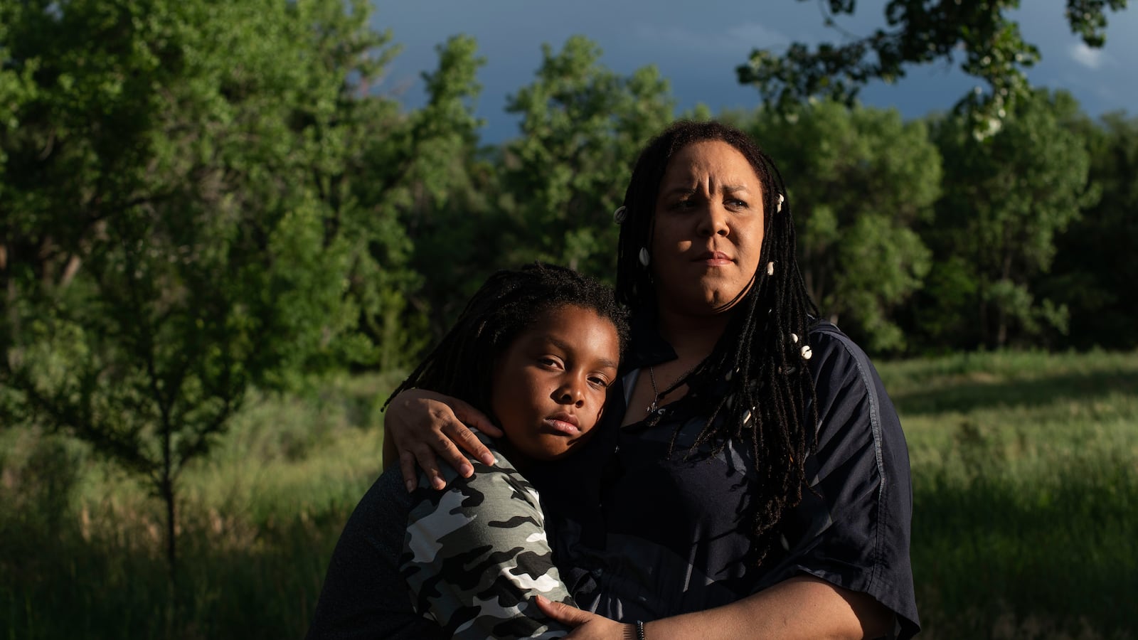 A mother and her 11-year-old son embrace in a park. They are both looking at the camera with serious expressions on their faces.