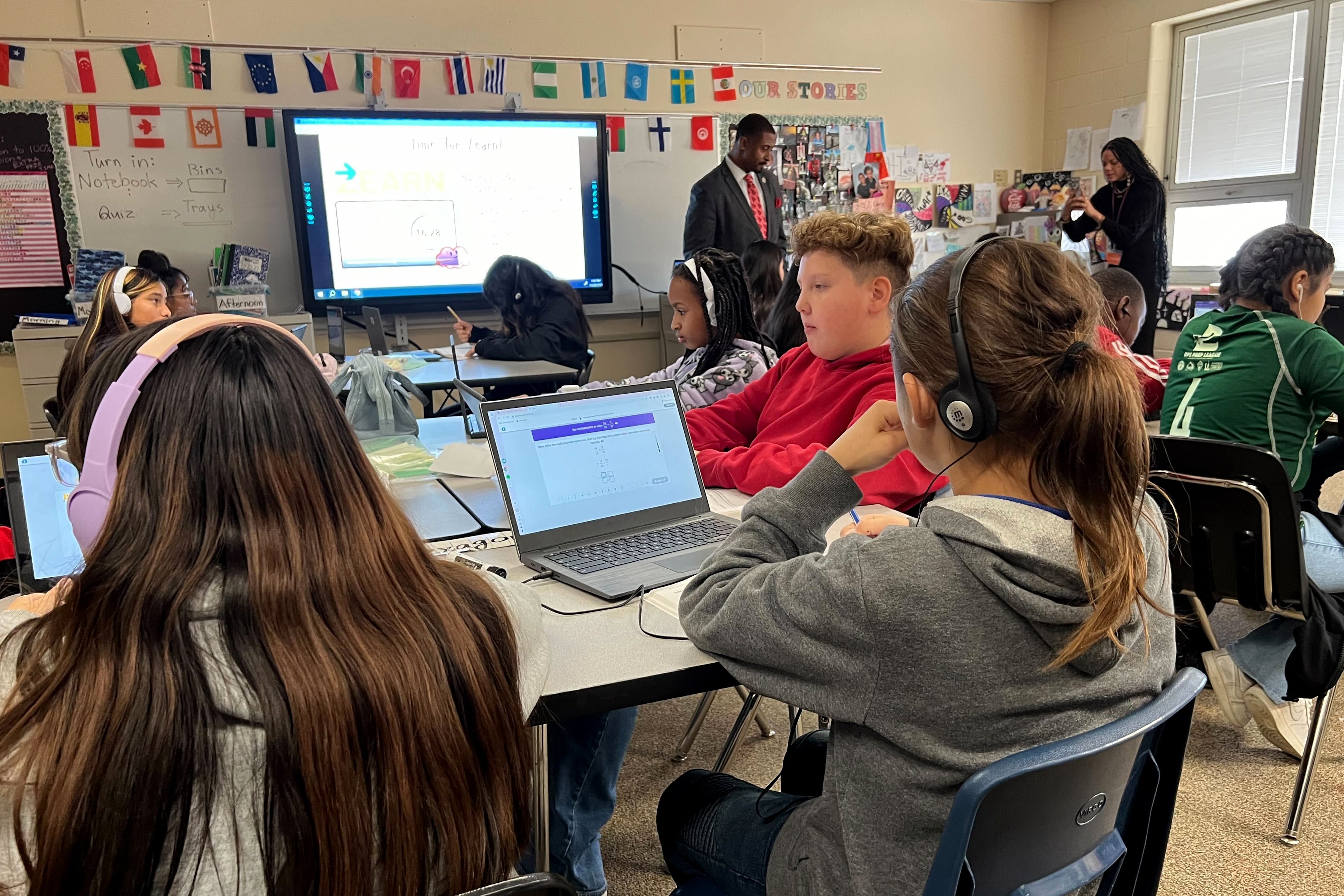 A class full of young students work from their desks while a couple of adults stand near the back of the class in the background.