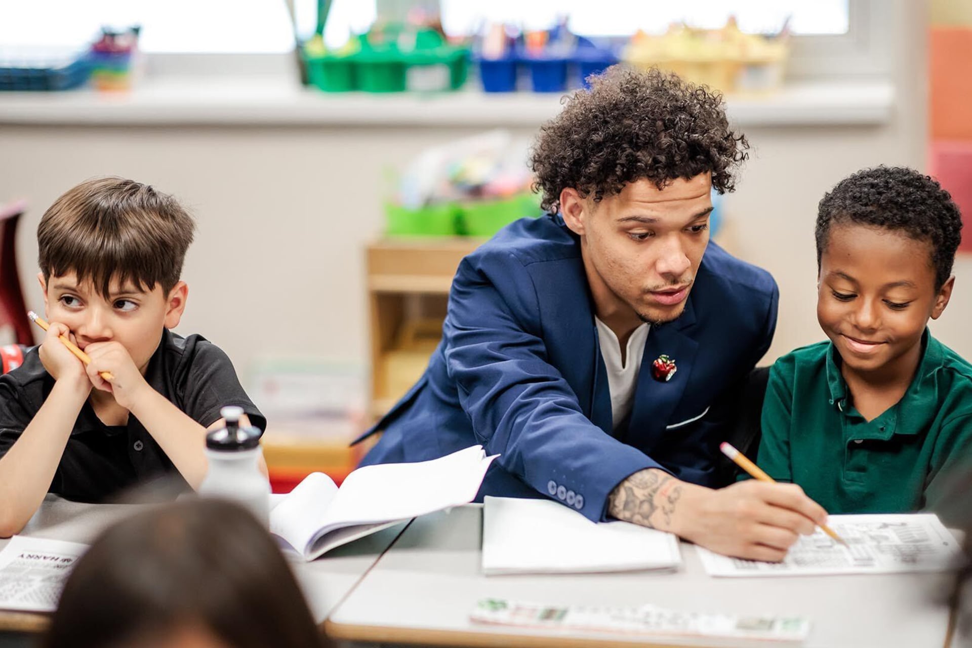 A young man with dark curly hair and wearing a blue suit, sits in between two young students at a table with a classroom setting in the background.